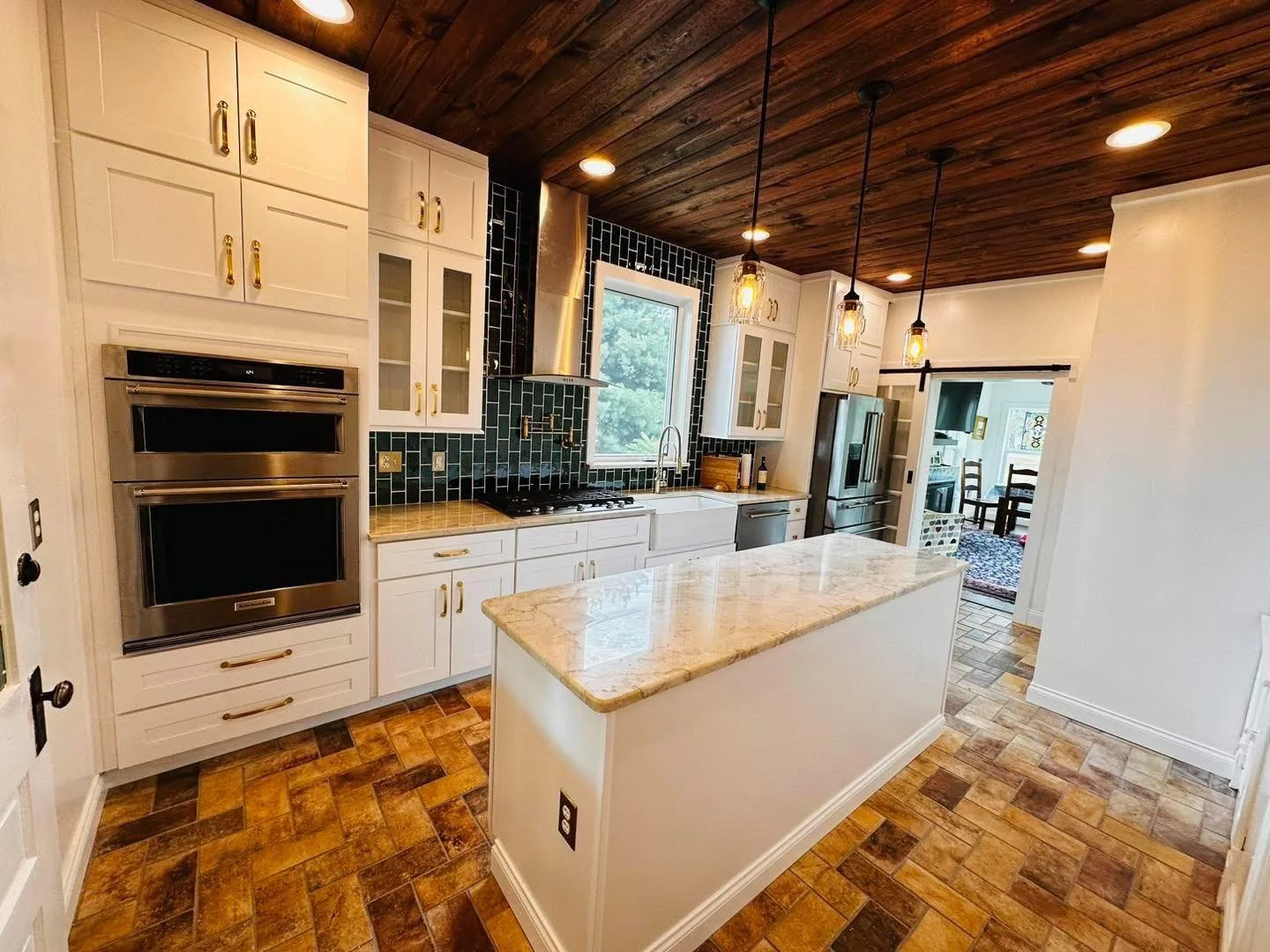 Modern kitchen with white cabinets and gold handles, a central island with beige marble countertop, a black tiled backsplash, stainless steel appliances, and a wooden ceiling with pendant lights.