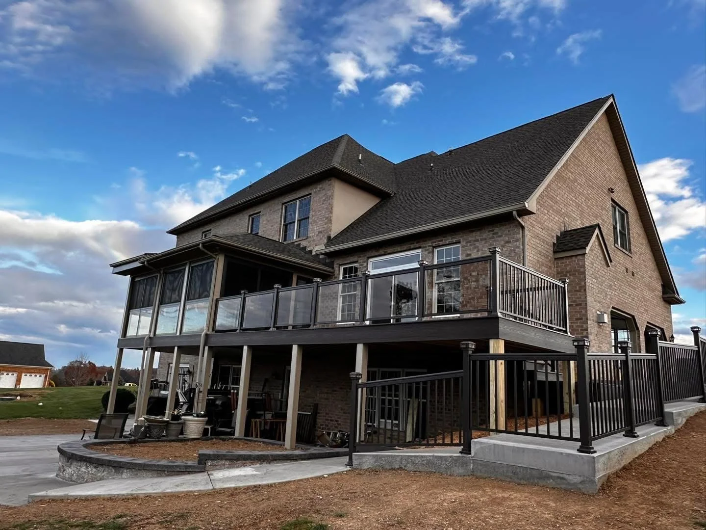 A large brick house with a multi-level deck and a fenced patio area. The sky is partly cloudy with blue and white clouds.