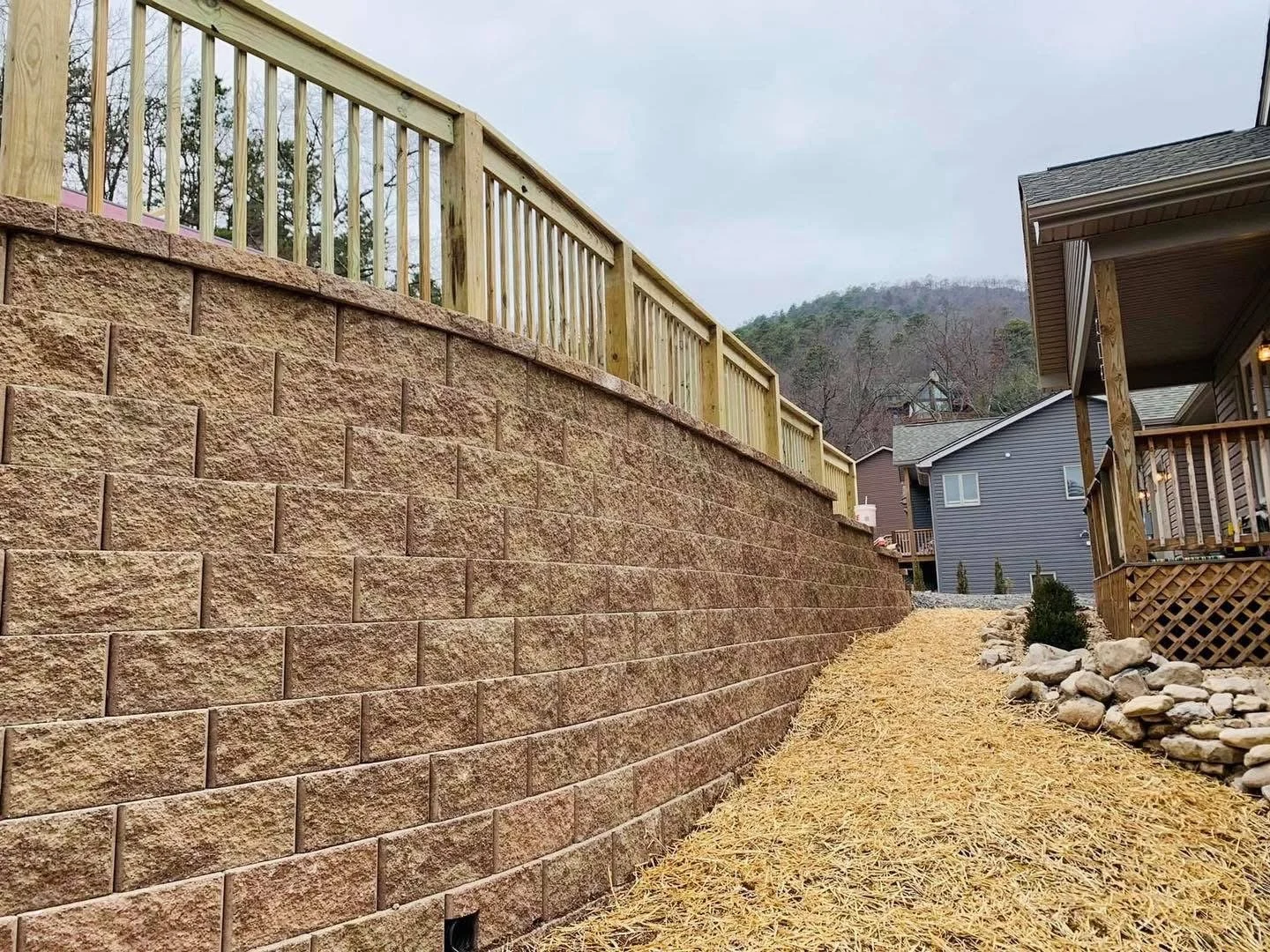 A backyard with a tall reddish-brown brick retaining wall topped by a new wooden fence, a gravel and straw-covered pathway, and houses in the background, with hills and trees.