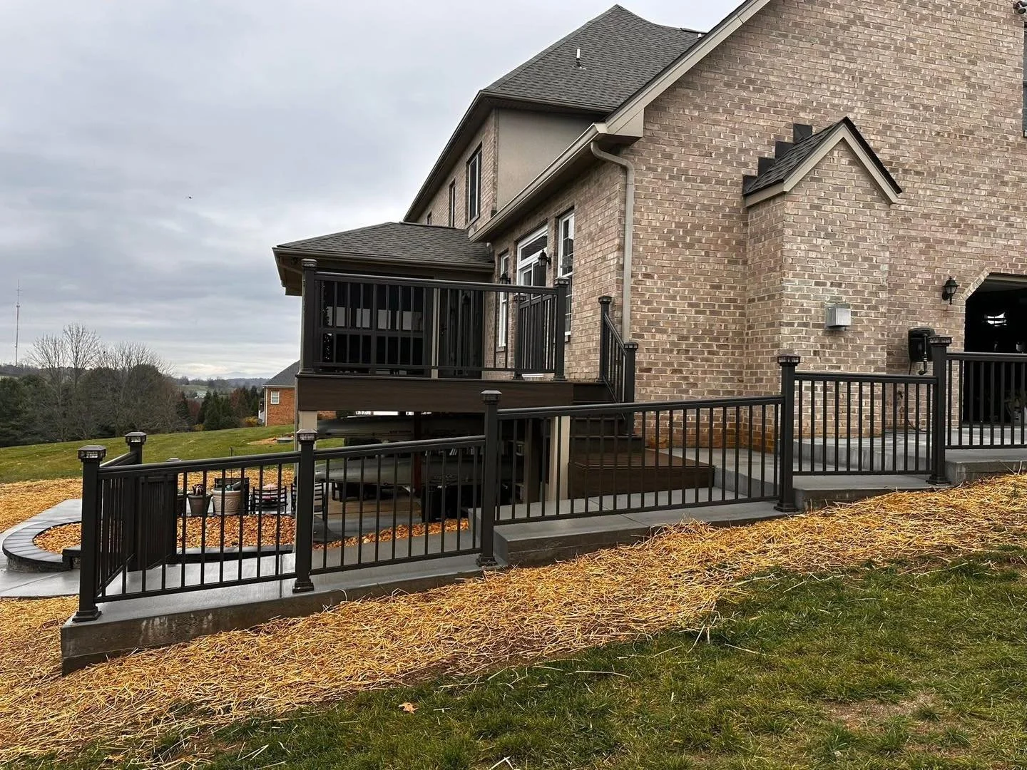 Backyard view of a two-story brick house with a wooden deck and black metal railing. The yard has grass and some autumn leaves, with a cloudy sky overhead.