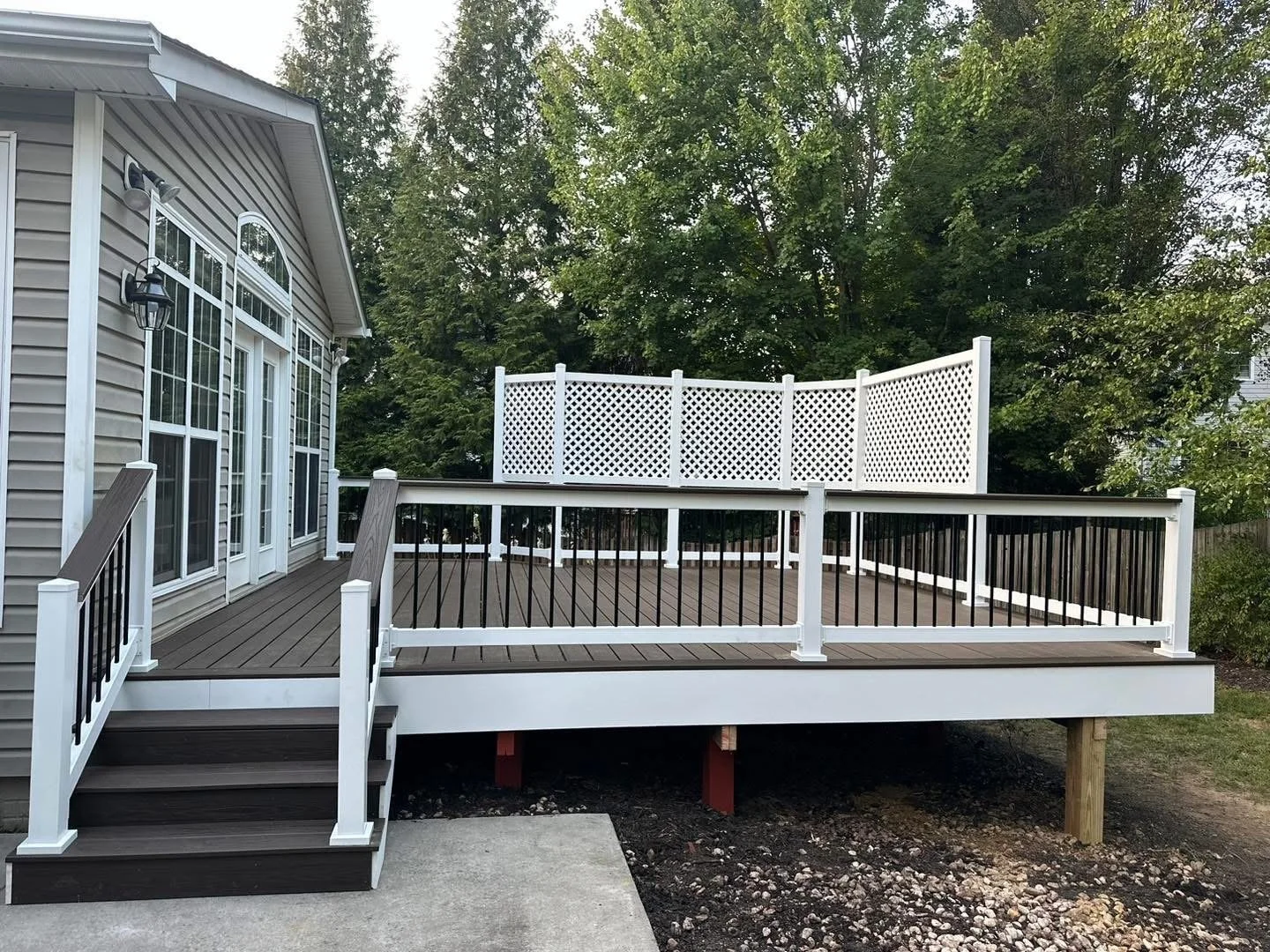Newly built wooden deck attached to a house with beige siding, surrounded by white and black railings, and a white lattice privacy screen, with a backdrop of green trees.