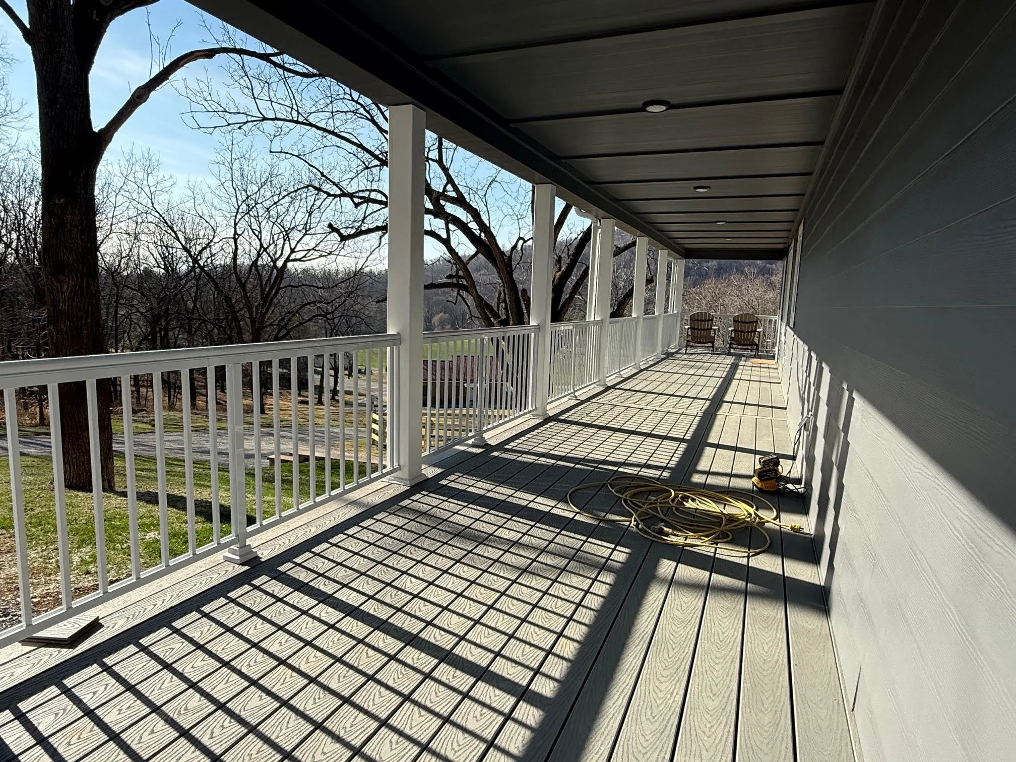 Empty porch with white railing, two chairs at the far end, and a coil of yellow extension cord on the wooden floor. Shadows cast by railing create a grid pattern on the floor. Trees and landscape visible in the background.