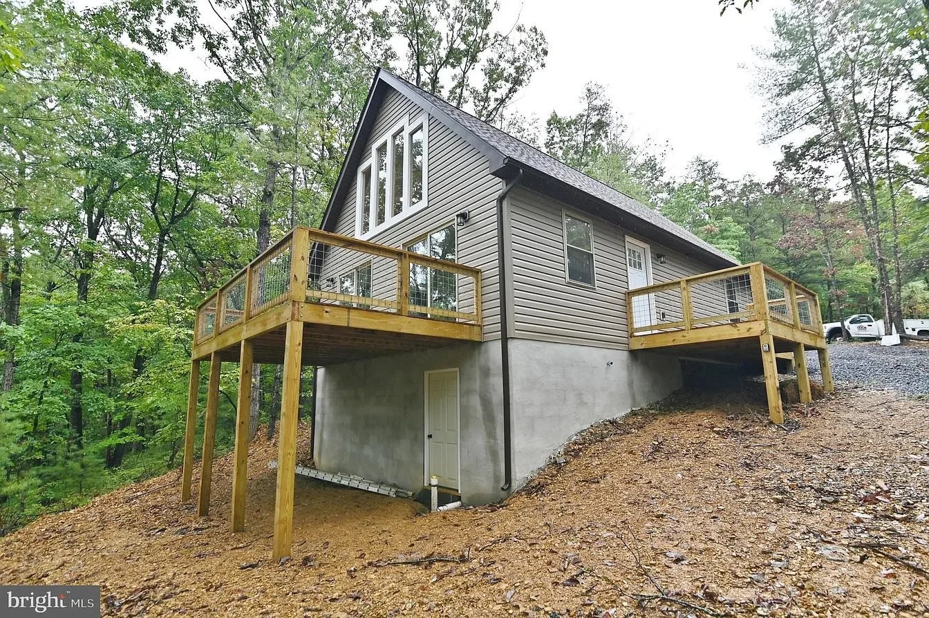 A two-story house with gray siding and a dark roof, with a large window on the upper floor. The house is built on a sloped terrain with a concrete foundation. There are two wooden decks with railings, one on each side of the house, supported by woode