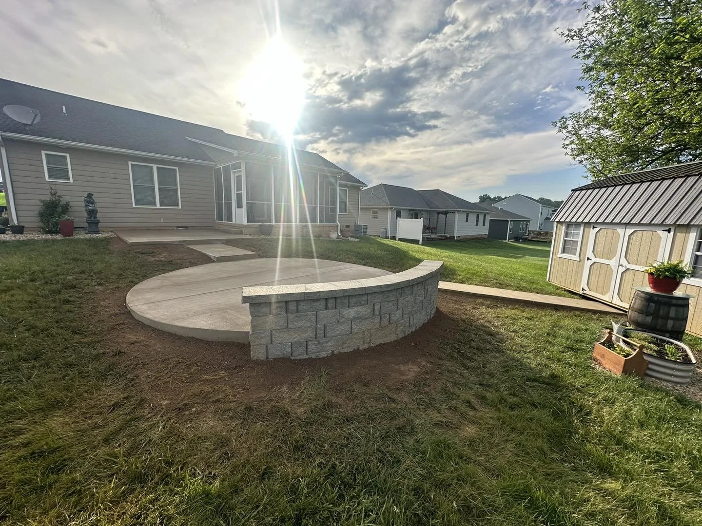 Backyard with a concrete patio, stone wall, and surrounding grass, with a house in the background under a partly cloudy sky.