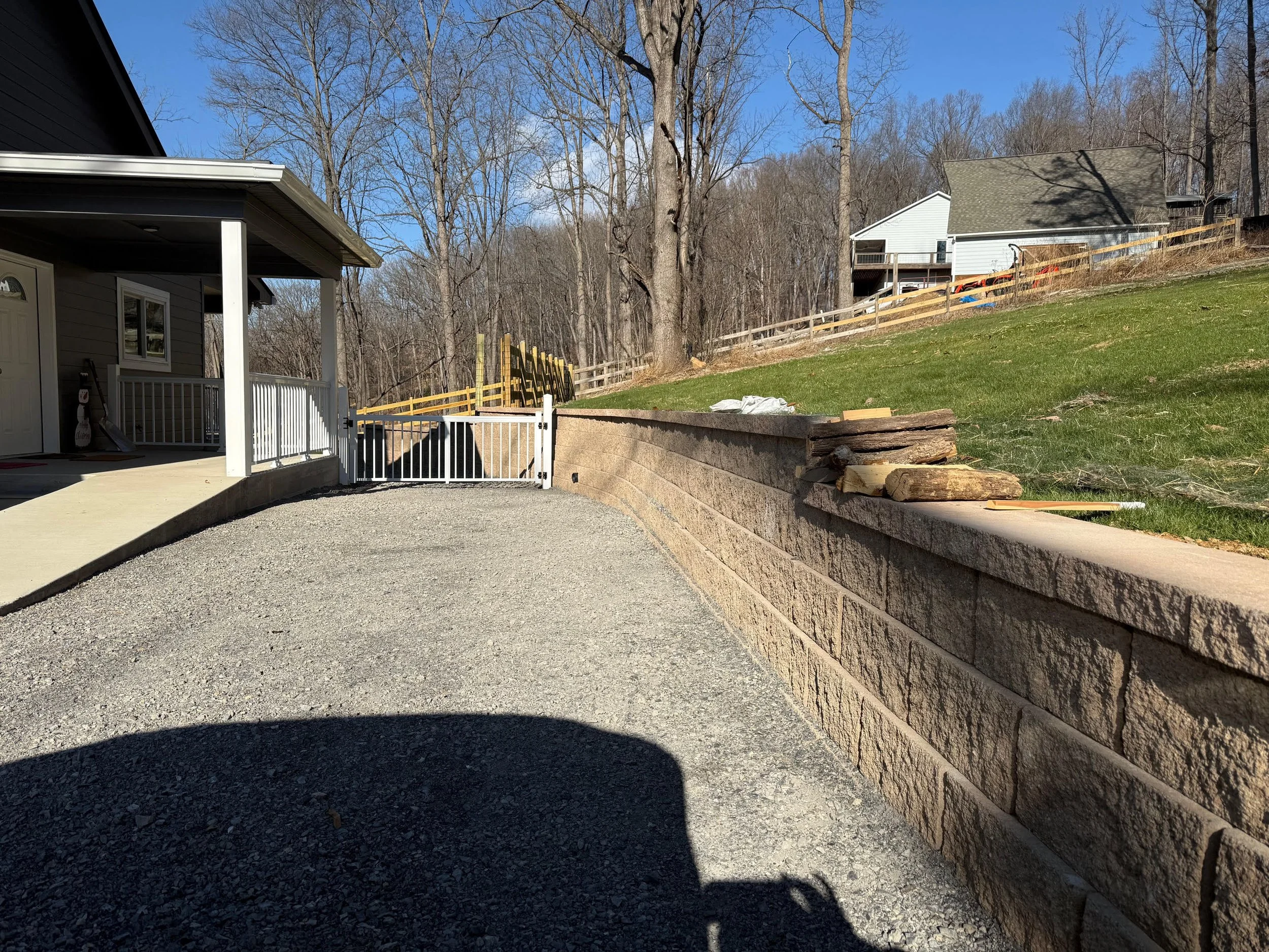 A backyard with a gravel driveway, a retaining wall made of large stone blocks, and a grassy hill with trees in the background. There's a small white gate at the end of the driveway and a house with a porch on the left. The sky is clear and sunny.