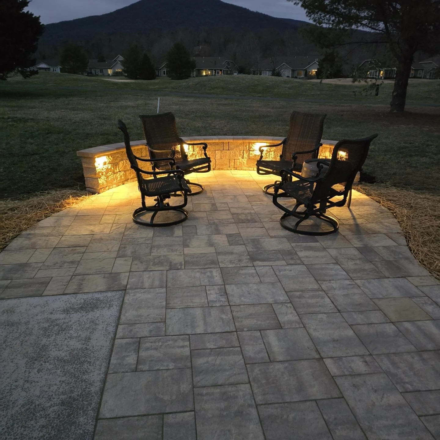 A cozy outdoor sitting area with four black chairs arranged in a semi-circle on a stone patio, illuminated by warm yellow lighting from beneath a stone wall, with a grassy yard, trees, residential houses, and hills in the background during dusk.