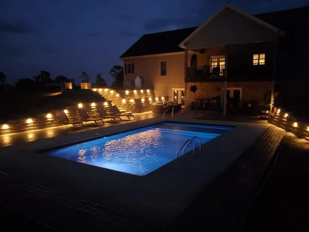 Nighttime scene of a backyard with a lit swimming pool, surrounded by lounge chairs and illuminated steps leading up to a house with a balcony.