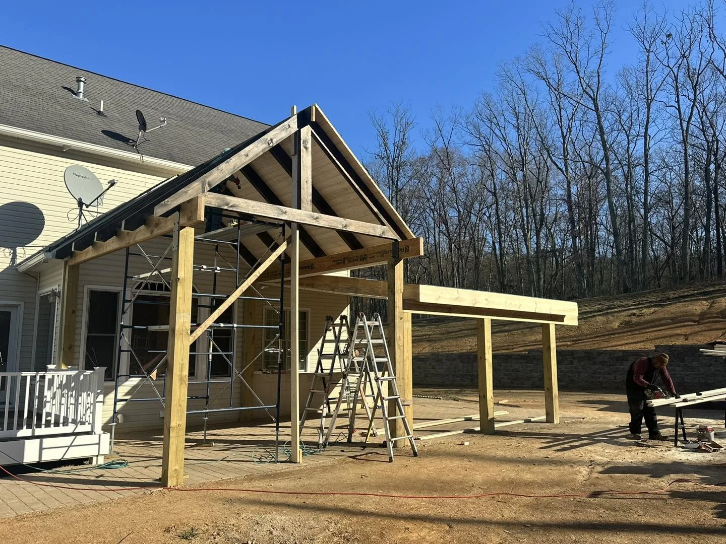 Construction of a backyard deck with a new wooden framework attached to a house. A worker is measuring or cutting wood, and ladders are present on site. The scene is set on a clear, sunny day with a wooded hill in the background.