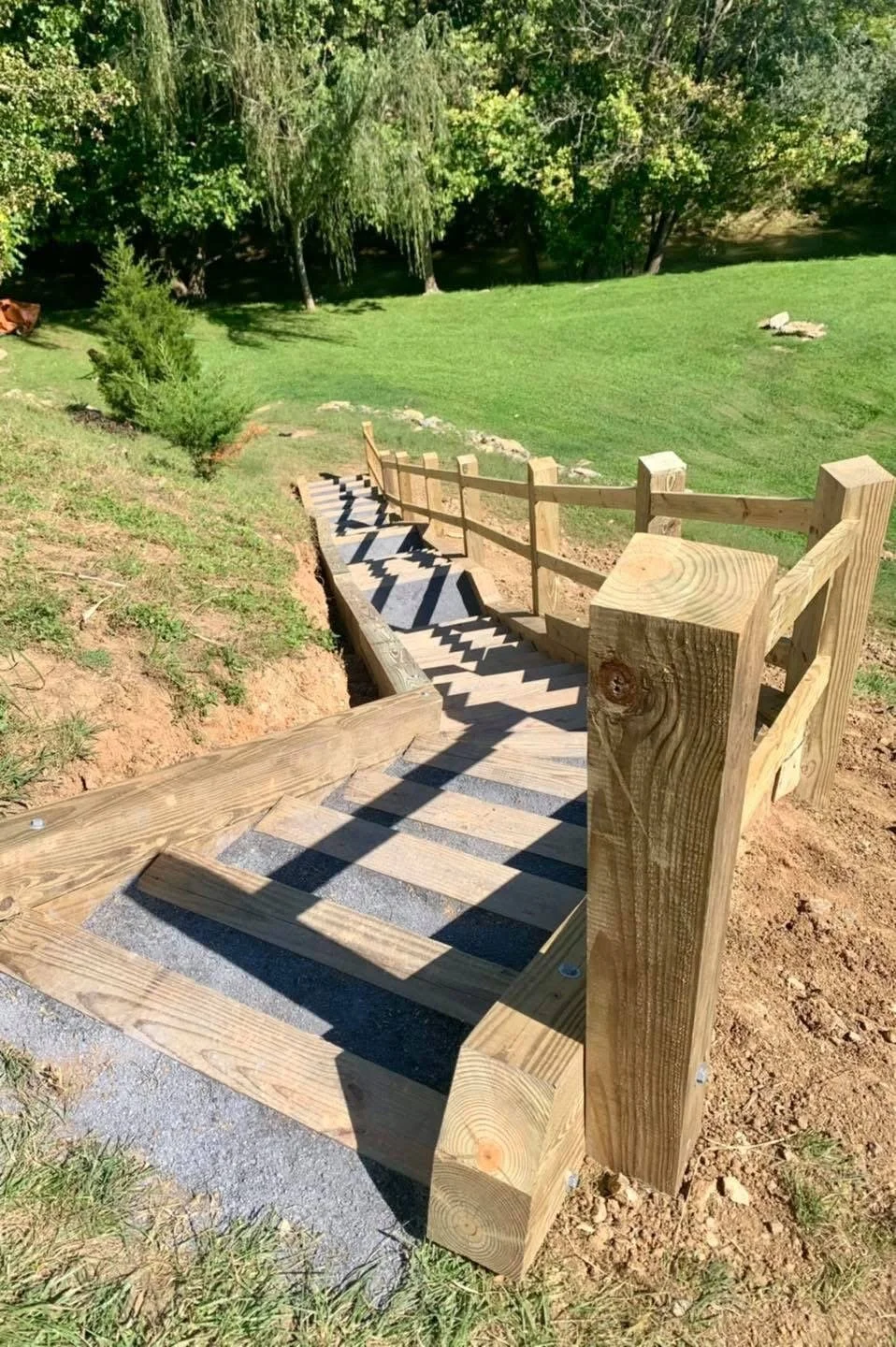 Newly built wooden staircase outdoors on a grassy hill, with shadows cast by the railing, leading down into a green lawn surrounded by trees.