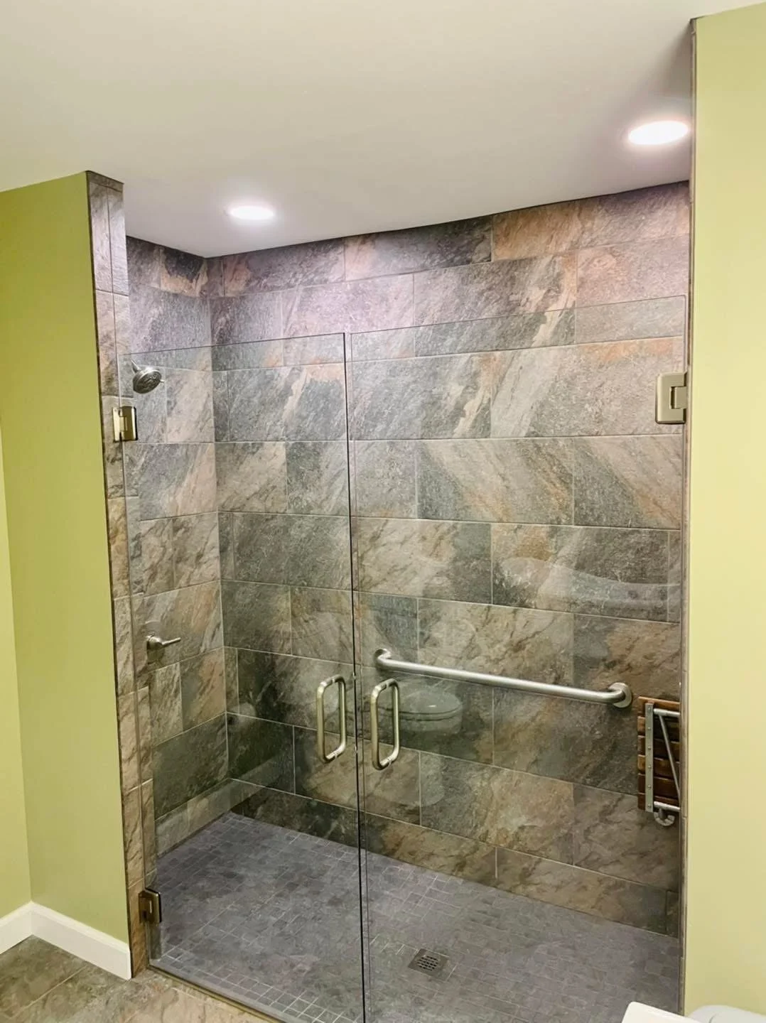 Walk-in shower with glass doors, large brown and gray stone tiles, a showerhead, a grab bar, and a small drain in the tiled floor.