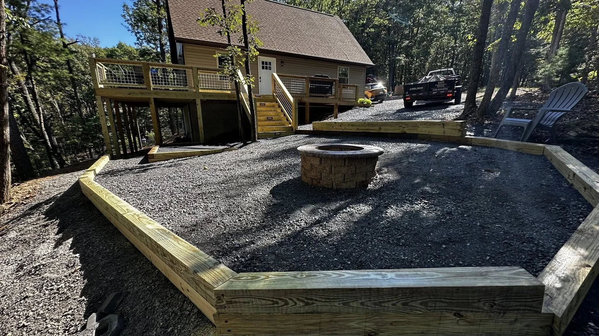 A backyard with a newly built wooden fire pit border and a fire pit in the center, with a house on a hill, a staircase, driveway, two parked vehicles, a couple of chairs, and trees surrounding the area.