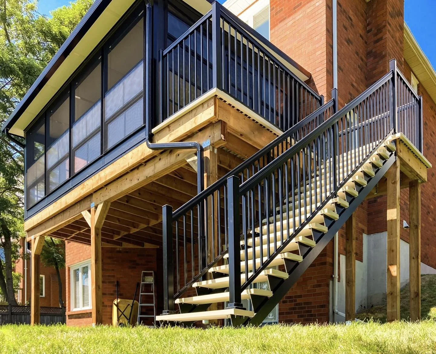 The image shows an outdoor wooden staircase with black railings leading up to a screened-in porch attached to a brick house. The porch has glass windows and is supported by wooden beams, with some tools and a ladder leaning against the house below.