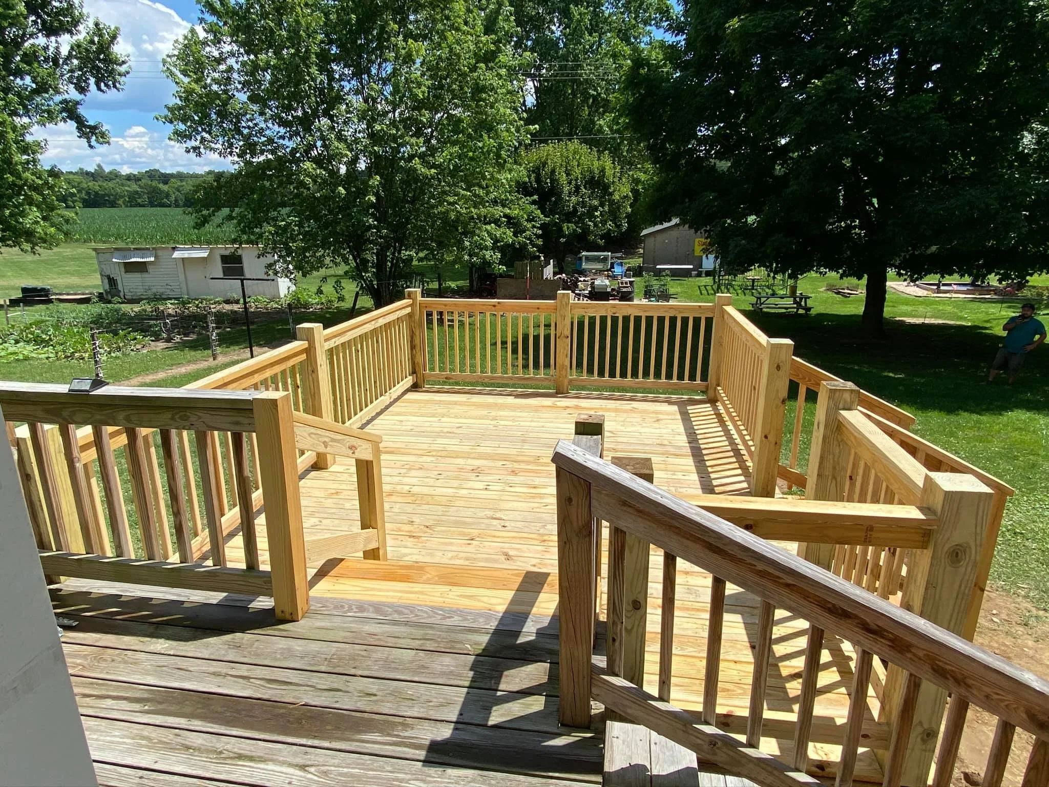 Newly constructed wooden deck with railings in a backyard, surrounded by trees and a green lawn, with a shed, picnic tables, and garden in the background.