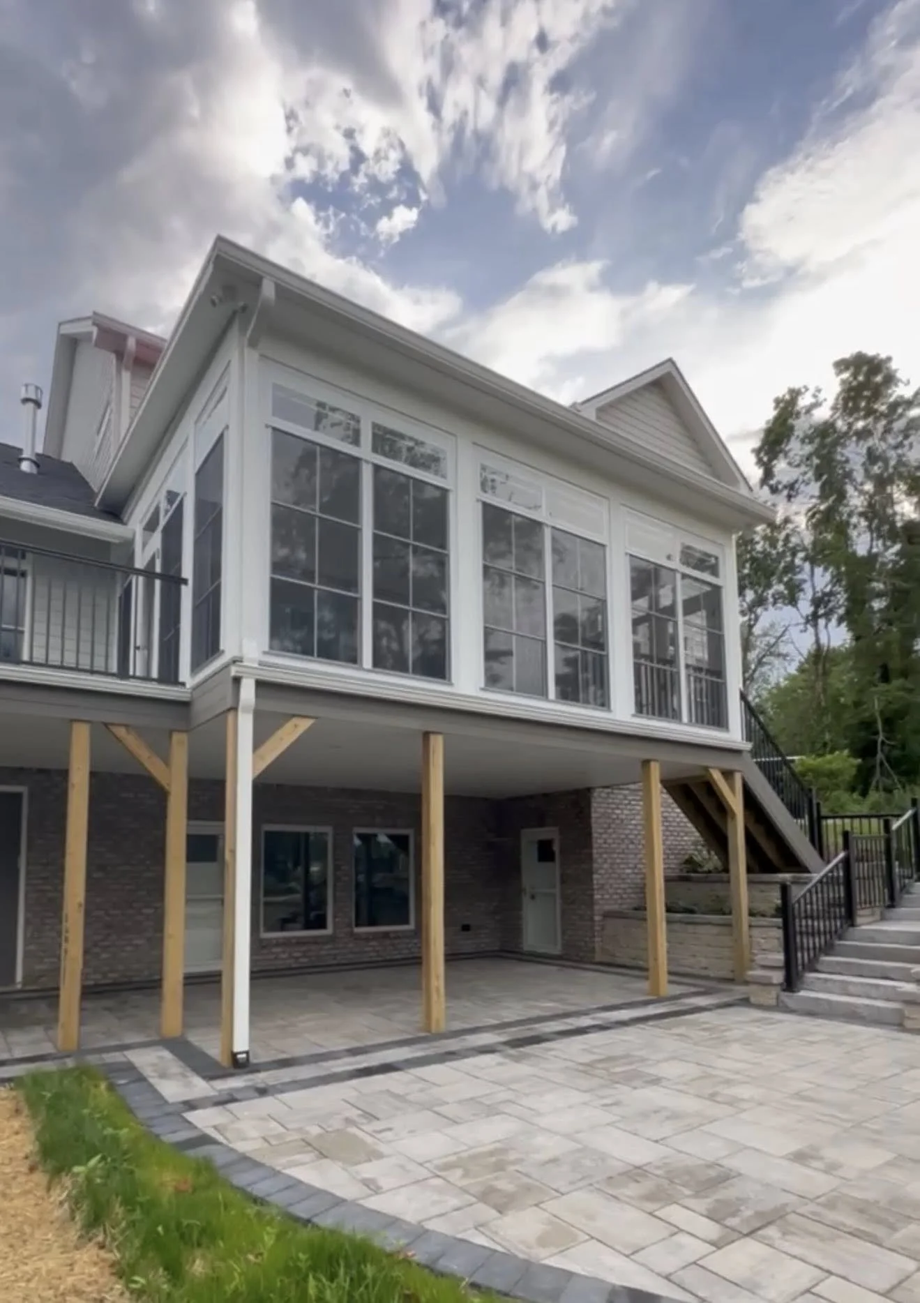 A house with a brick foundation and an elevated enclosed porch with large windows, supported by wooden posts, and stairs leading up to the entrance on the right side. The yard has a paved area and some grass, with trees in the background and a partly cloudy sky above.