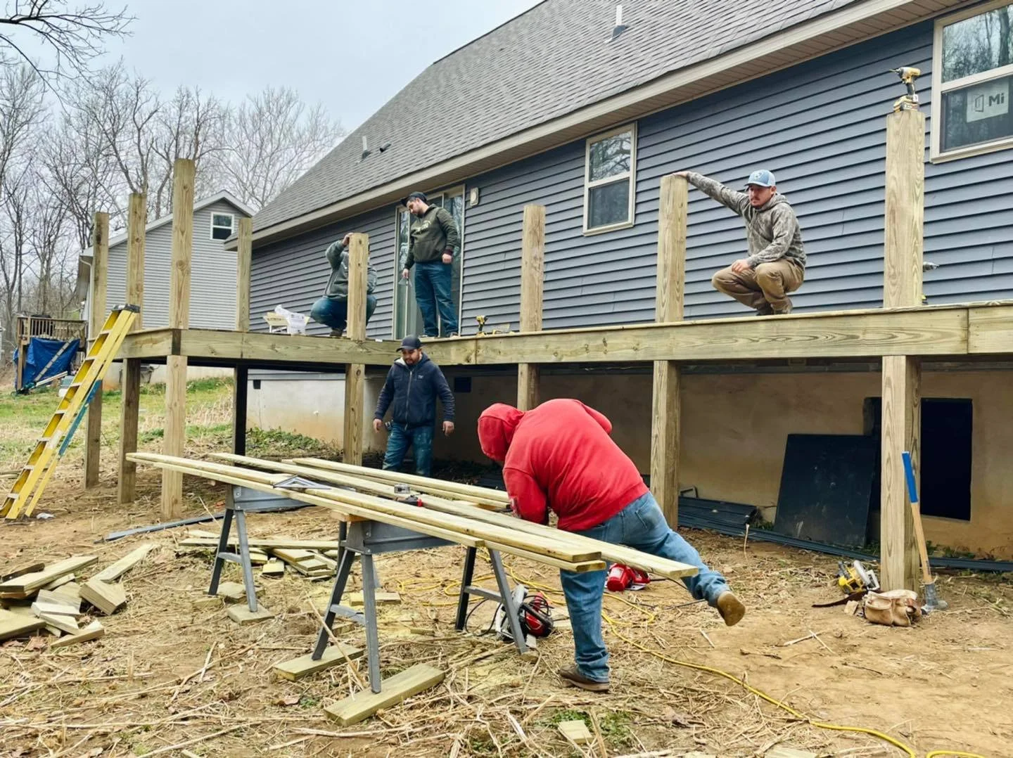 Group of people working on building a wooden deck outside a house, with tools and lumber scattered around.