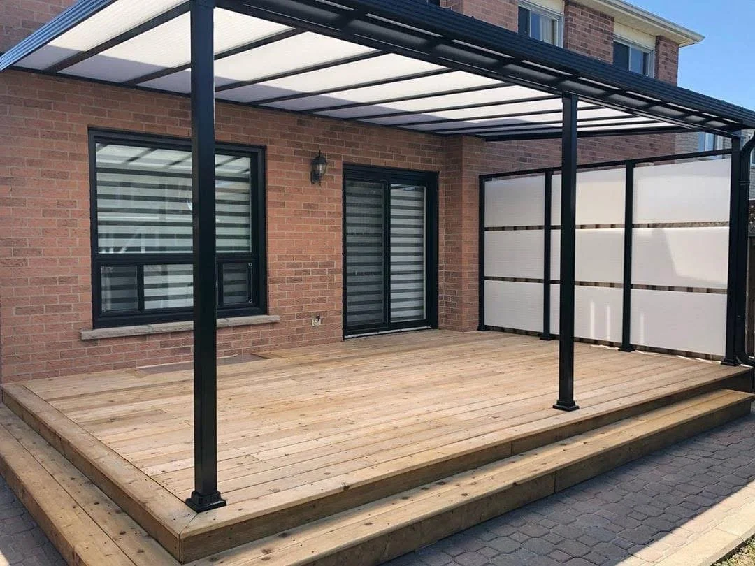 Wooden deck attached to a brick house with black-framed sliding glass doors, a window, and a black metal and white privacy panel fencing, partially covered by a black and white awning.