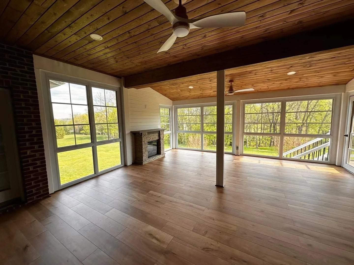 Unfurnished sunroom with large windows, wooden ceiling, hardwood floors, stone fireplace, ceiling fans, and views of green lawn and trees outside.
