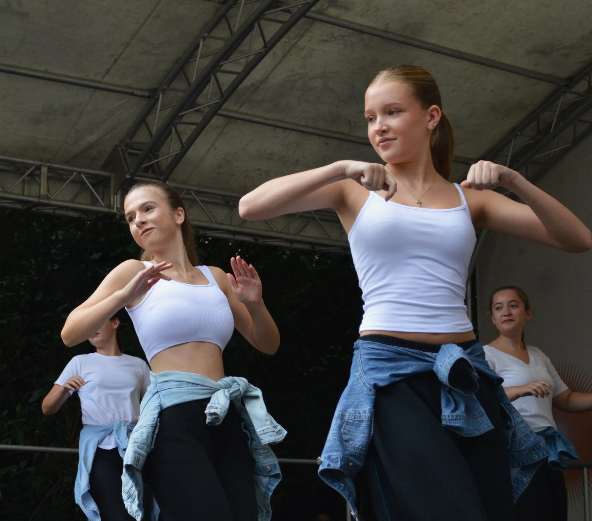 Vier junge Frauen tanzen auf einer Bühne, tragen weiße Oberteile und Jeansjacken um die Taille, während sie ihre Arme in verschiedenen Positionen bewegen.