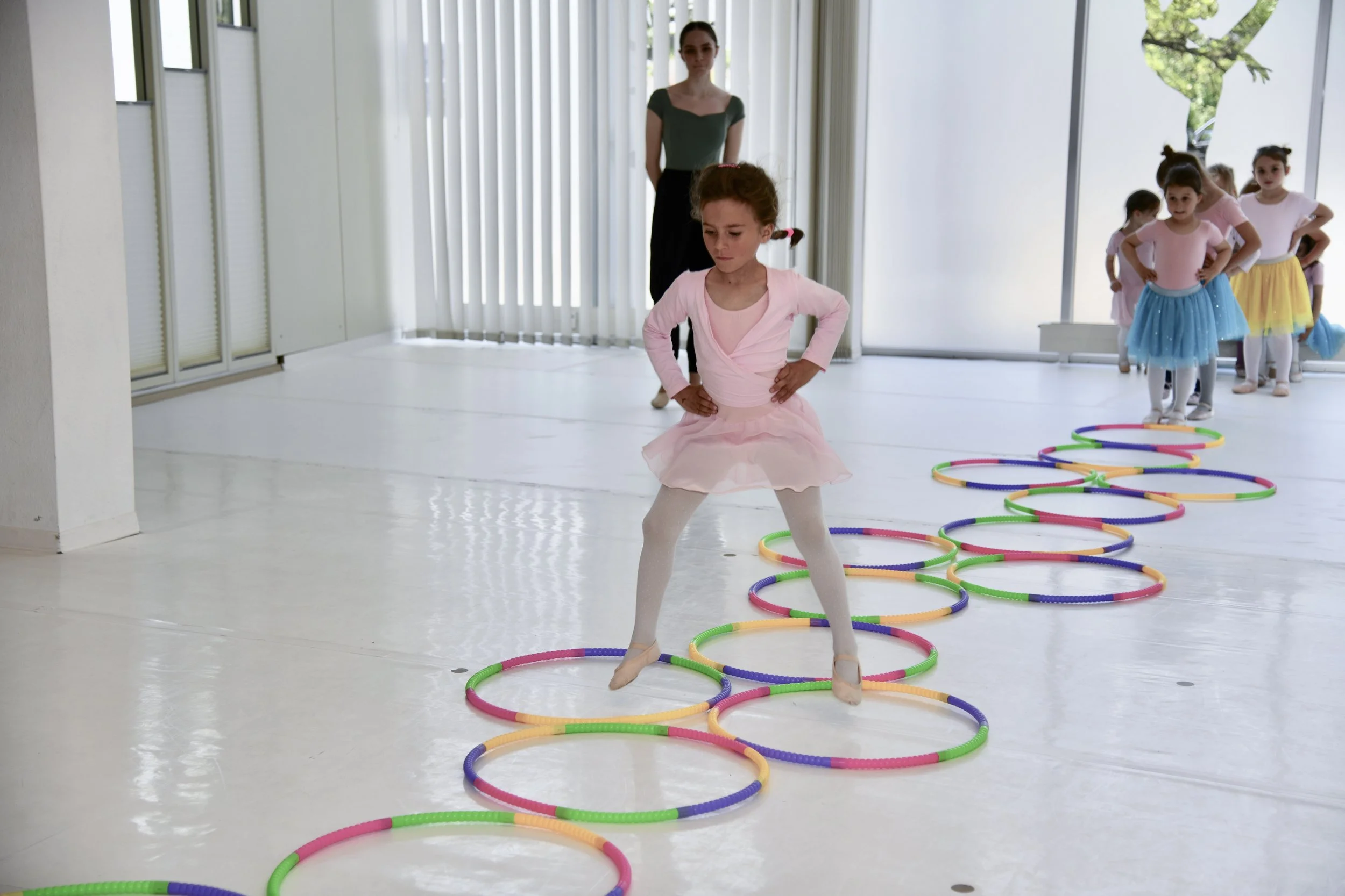 Tänzerin übt im Ballettunterricht, während andere Kinder im Hintergrund warten. Sie trägt einen pinken Tütü und tanzt auf einem रंगीला Hoop-Loop-Parcours.