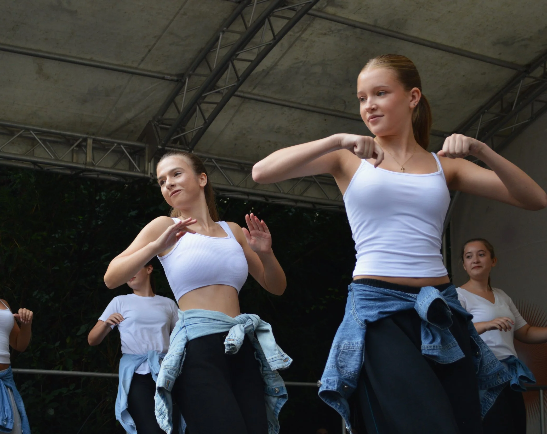 Jugendliche Mädchen tanzen auf einer Bühne, tragen weiße Tanktops, Jeansjacken um die Taille gebunden, beim Tanzen mit fröhlichen Gesichtsausdrücken.