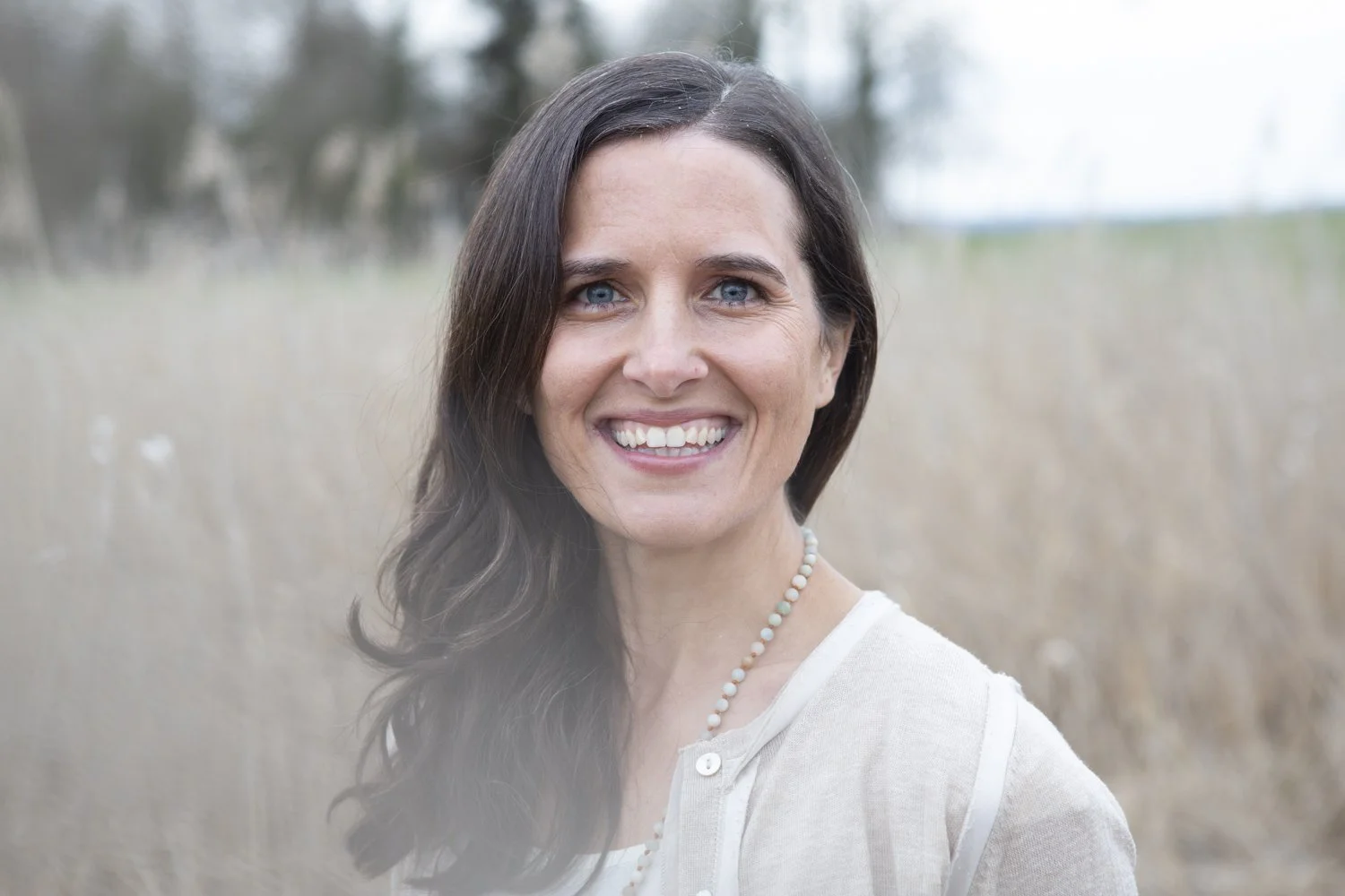 Lächelnde Frau mit dunklen Haaren, blaue Augen, träg einer Perlenkette und einem weißen Hemd im Freien im Feld.