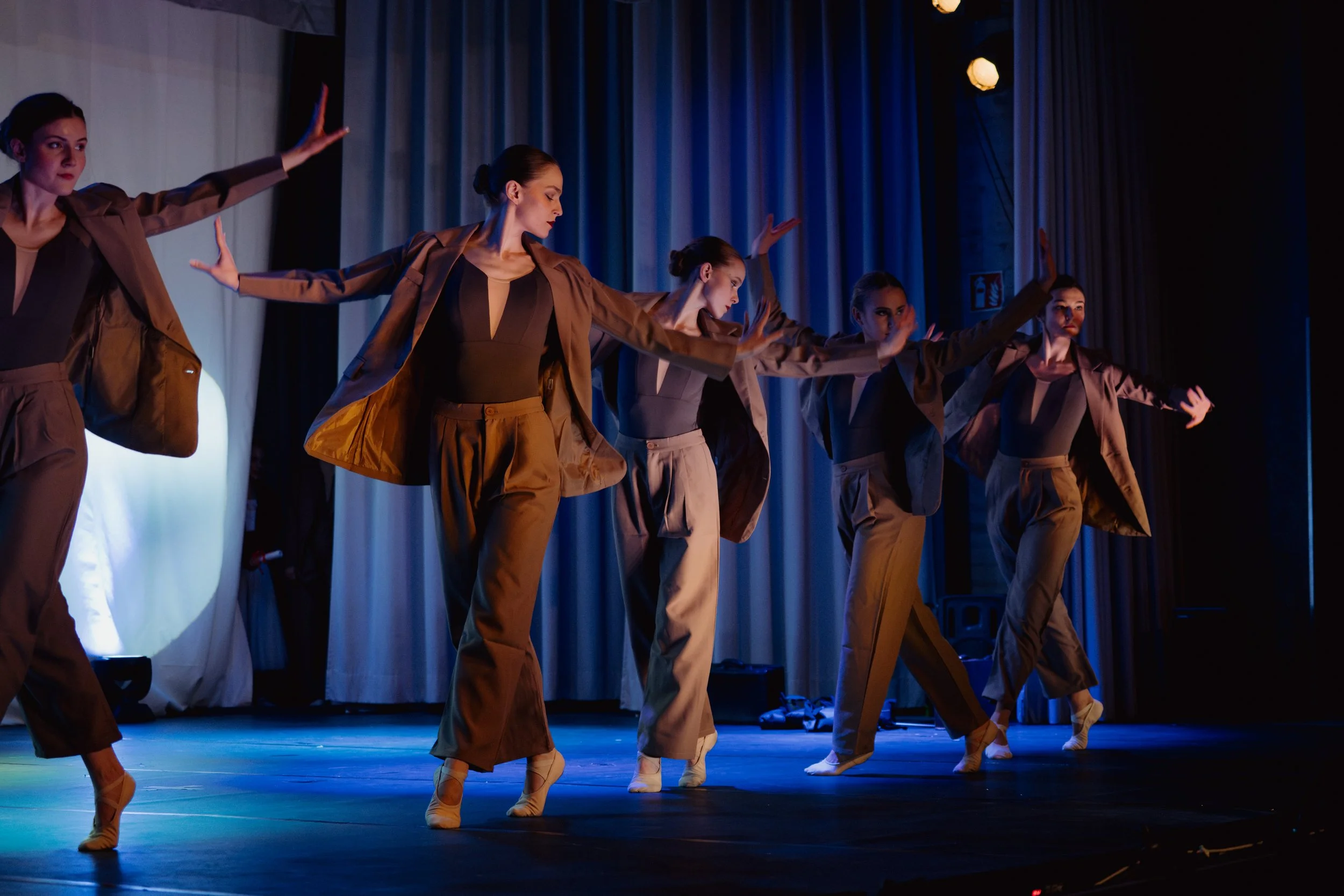 Tanzgruppe von sechs Frauen auf der Bühne, tanzen in braunen Outfits mit blauer Beleuchtung im Hintergrund.