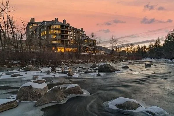 A modern residential building beside a river with rocks, during sunset or sunrise, with a partly cloudy sky and bare trees in the scene.