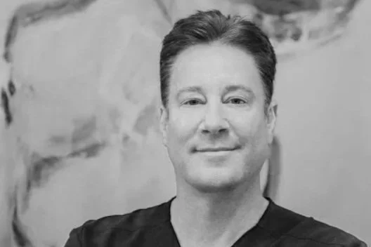 Black-and-white portrait of Mark M. Hamilton, MD, smiling slightly at the camera, wearing a dark shirt, standing in front of a blurred landscape background.