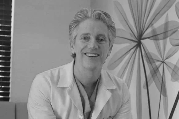 A smiling John J. Martin, Jr.,MD, wearing a white lab coat, sitting in front of a wall with a large, leafy plant in the background.