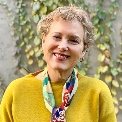 A middle-aged woman with short, curly hair smiling outdoors in front of a wooden fence covered with green and yellow ivy. She is wearing a yellow sweater and a colorful patterned scarf.
