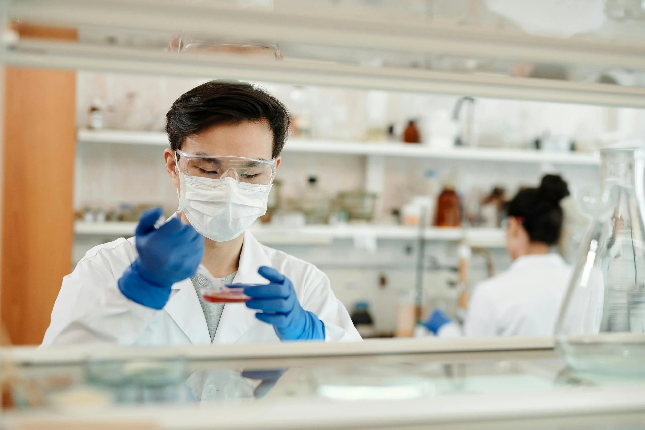 A scientist wearing safety goggles, a face mask, and blue gloves is working in a laboratory, holding a petri dish and examining its contents. Another scientist is visible in the background, also working in the lab.