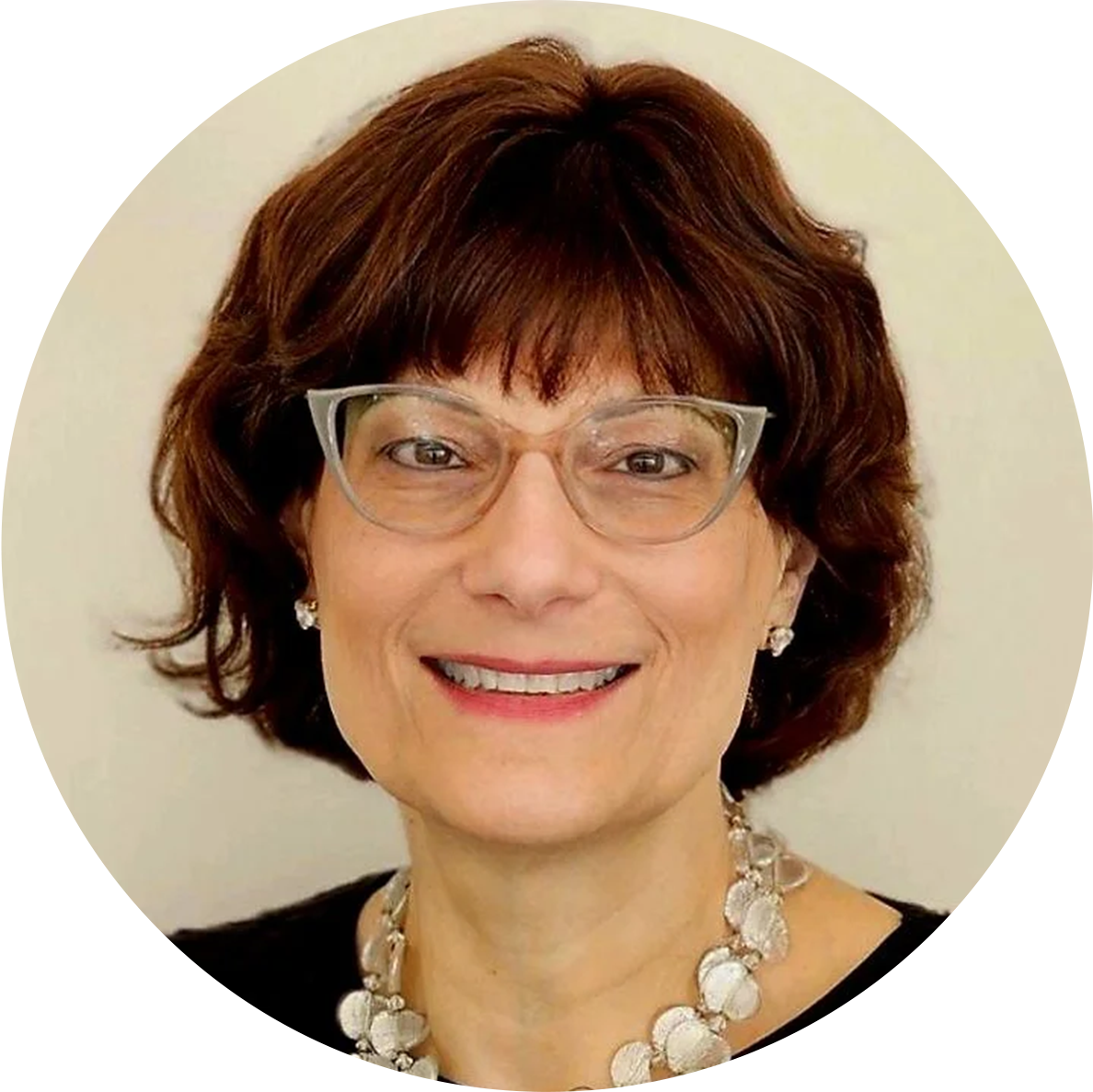 A middle-aged woman with short brown hair, wearing glasses, pearl earrings, a pearl necklace, and a black top, smiling against a plain light-colored background.