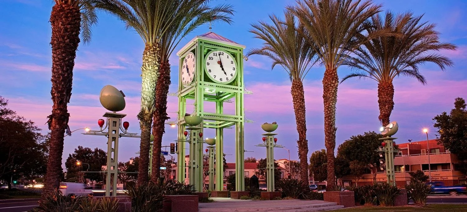 A clock tower with a green structure, surrounded by tall palm trees, at sunset, with buildings and a street with moving cars in the background.