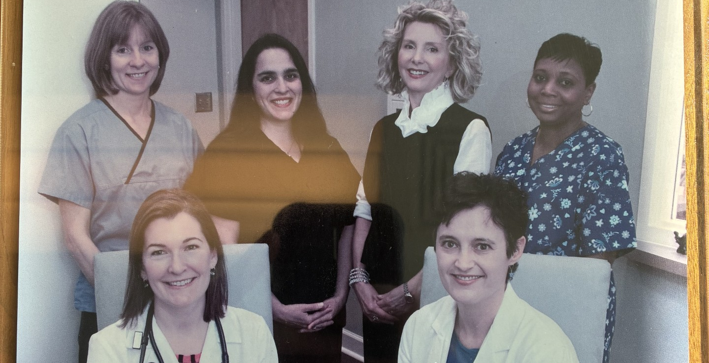 Group of seven women, some in medical attire, smiling in an office or hospital setting.