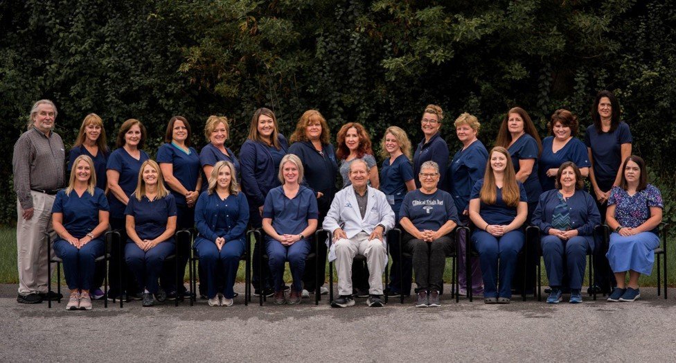 Group photo of healthcare professionals in scrubs and lab coats outdoors, with trees in the background.
