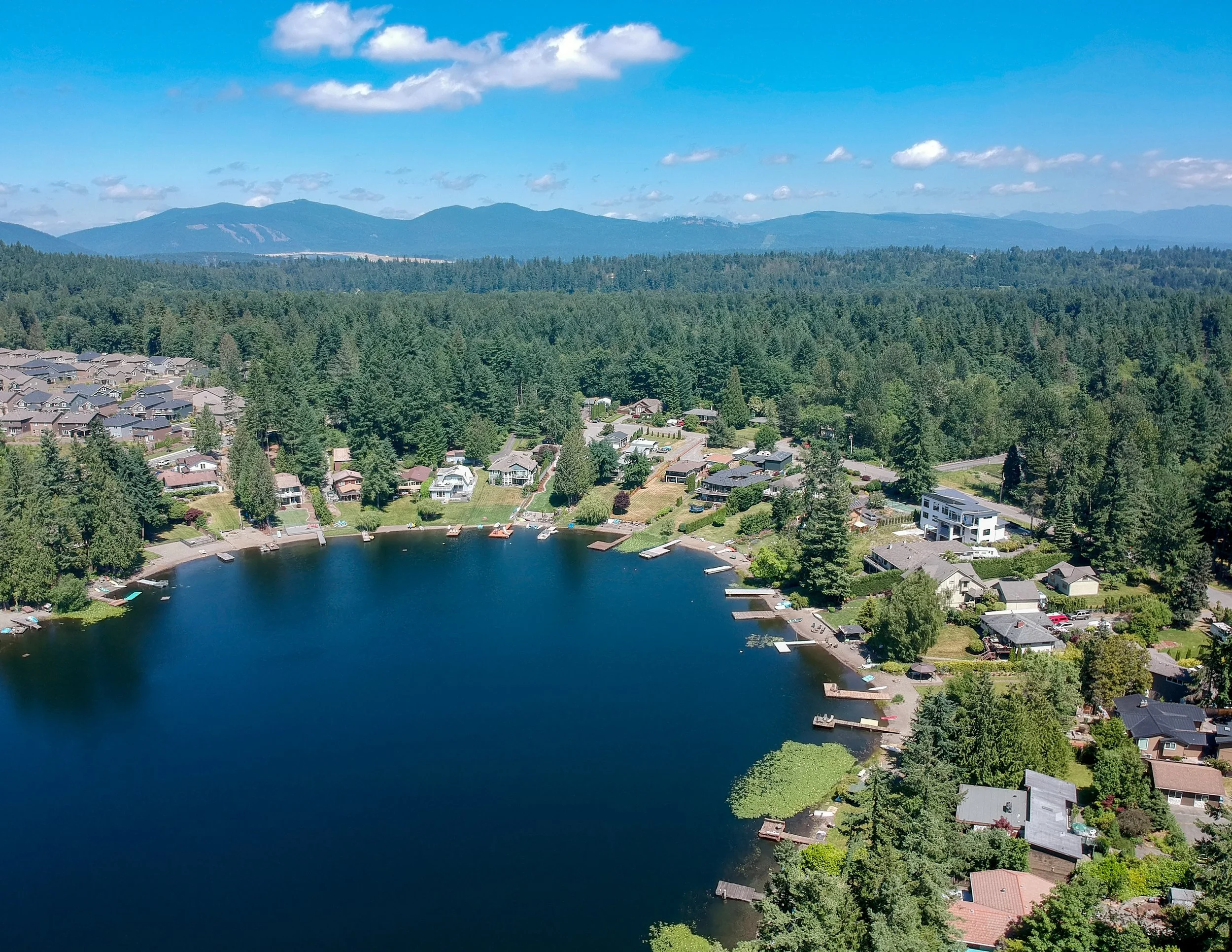 Aerial view of a residential neighborhood surrounding a small lake with boats, trees, and houses, with forested hills and mountains in the background on a clear, sunny day.