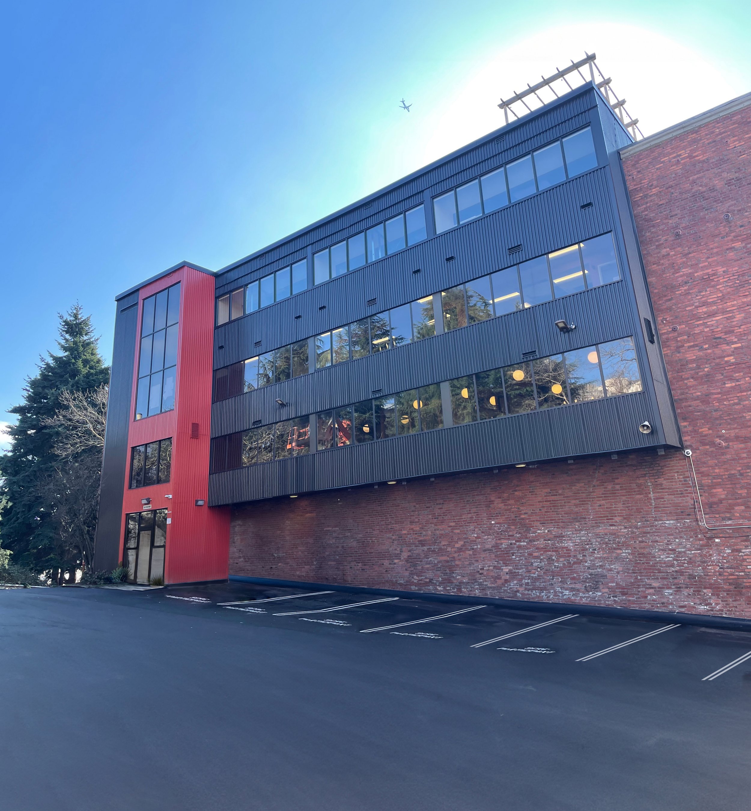 Modern multi-story building with a red section on the left and black siding, large reflective windows, brick wall base, parking lot in front, and an airplane flying overhead against a clear blue sky.