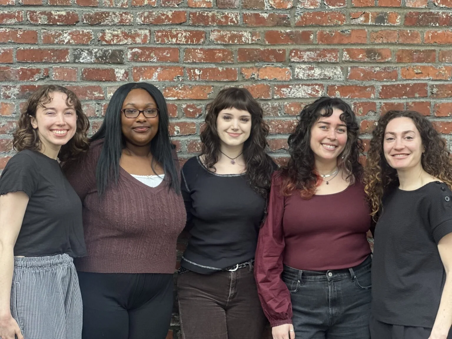 Five women standing in front of a brick wall, smiling at the camera.