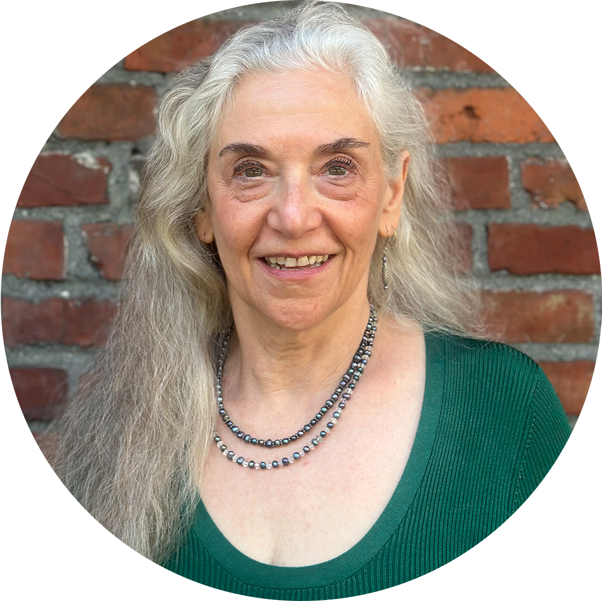 A smiling elderly woman with long, wavy gray hair, wearing a green top and layered pearl necklaces standing in front of a brick wall.