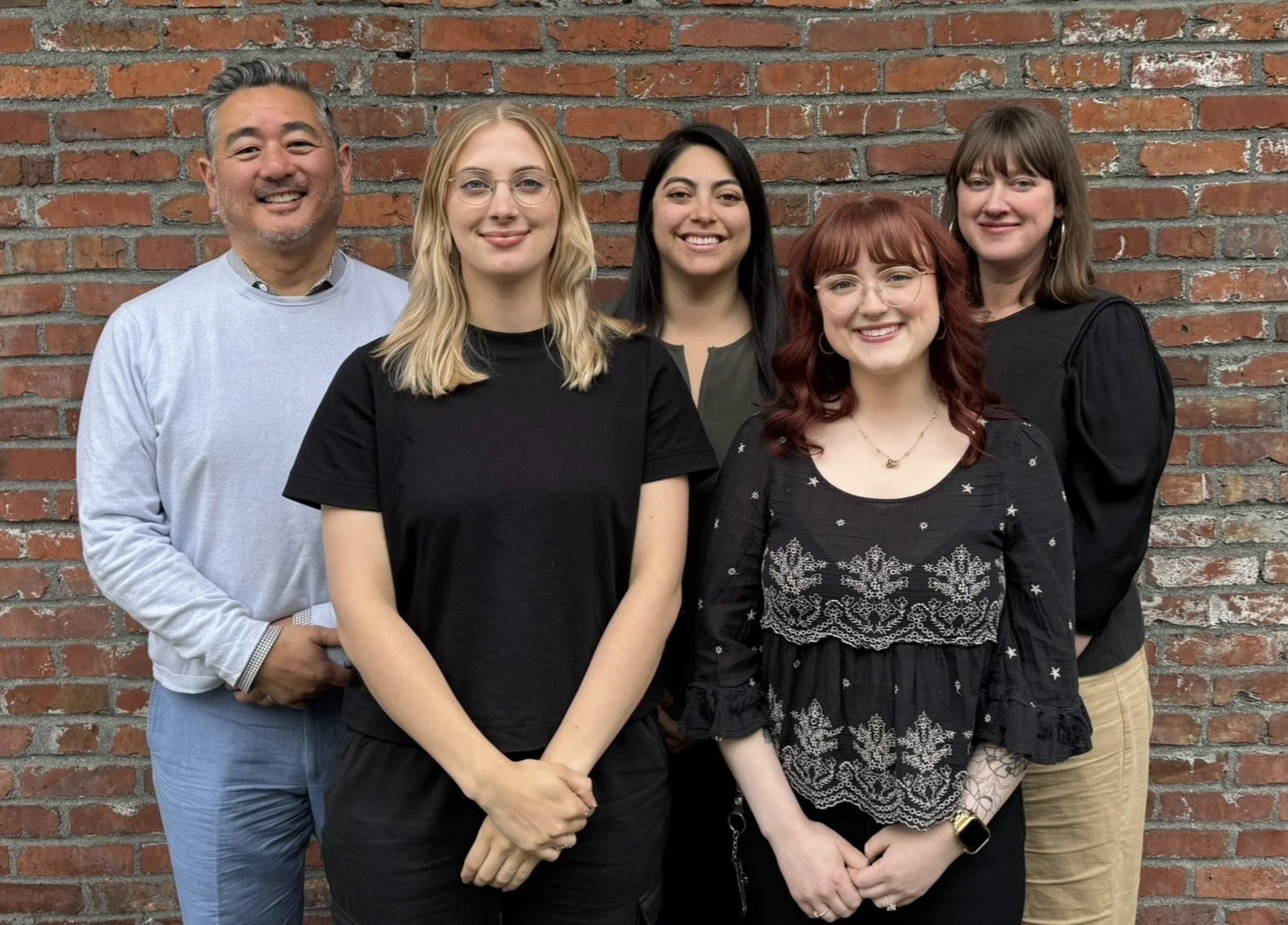 Group of five people standing in front of a brick wall, smiling at the camera.
