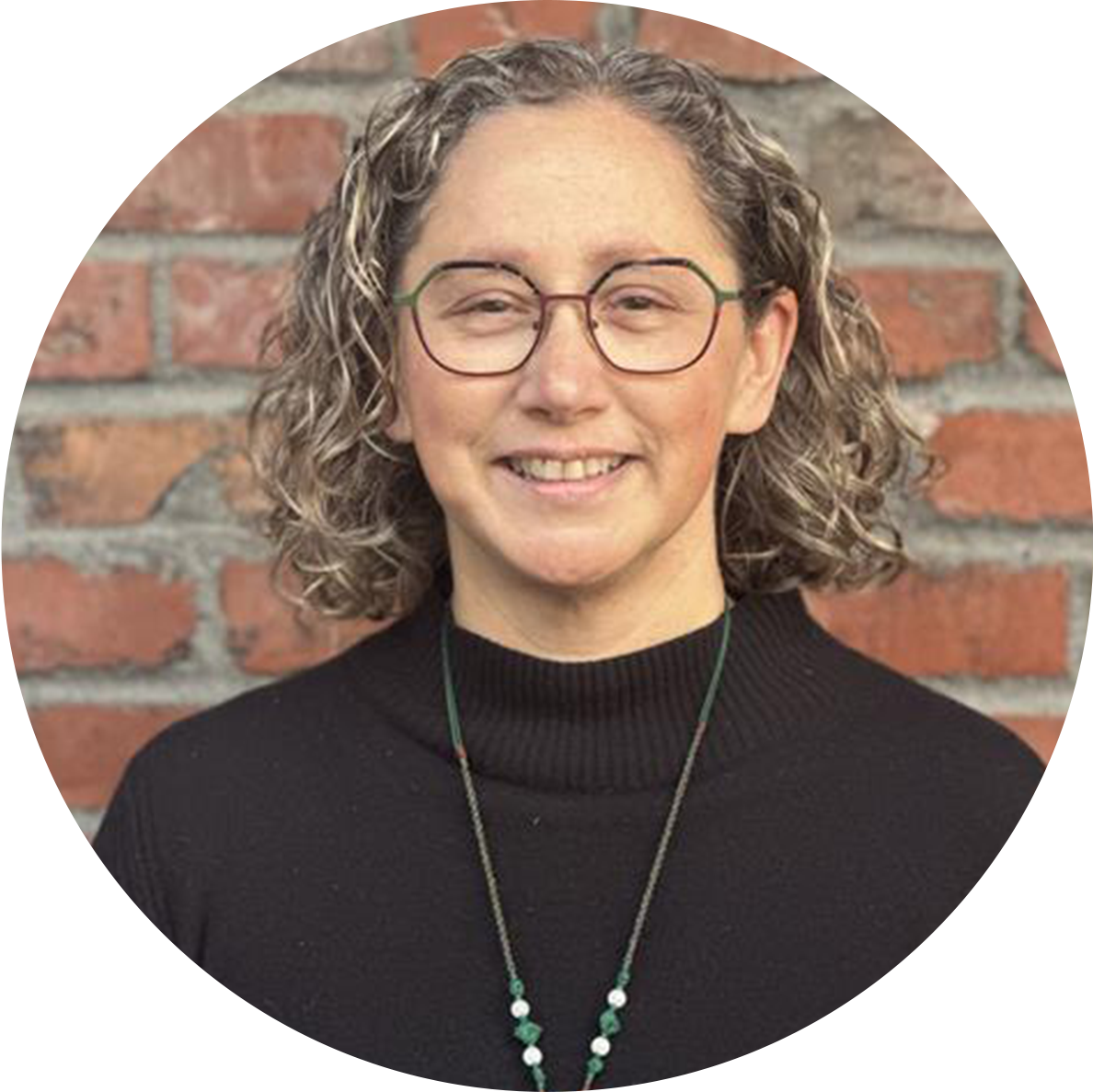 A woman with curly gray hair, glasses, and a black turtleneck sweater, smiling in front of a brick wall.