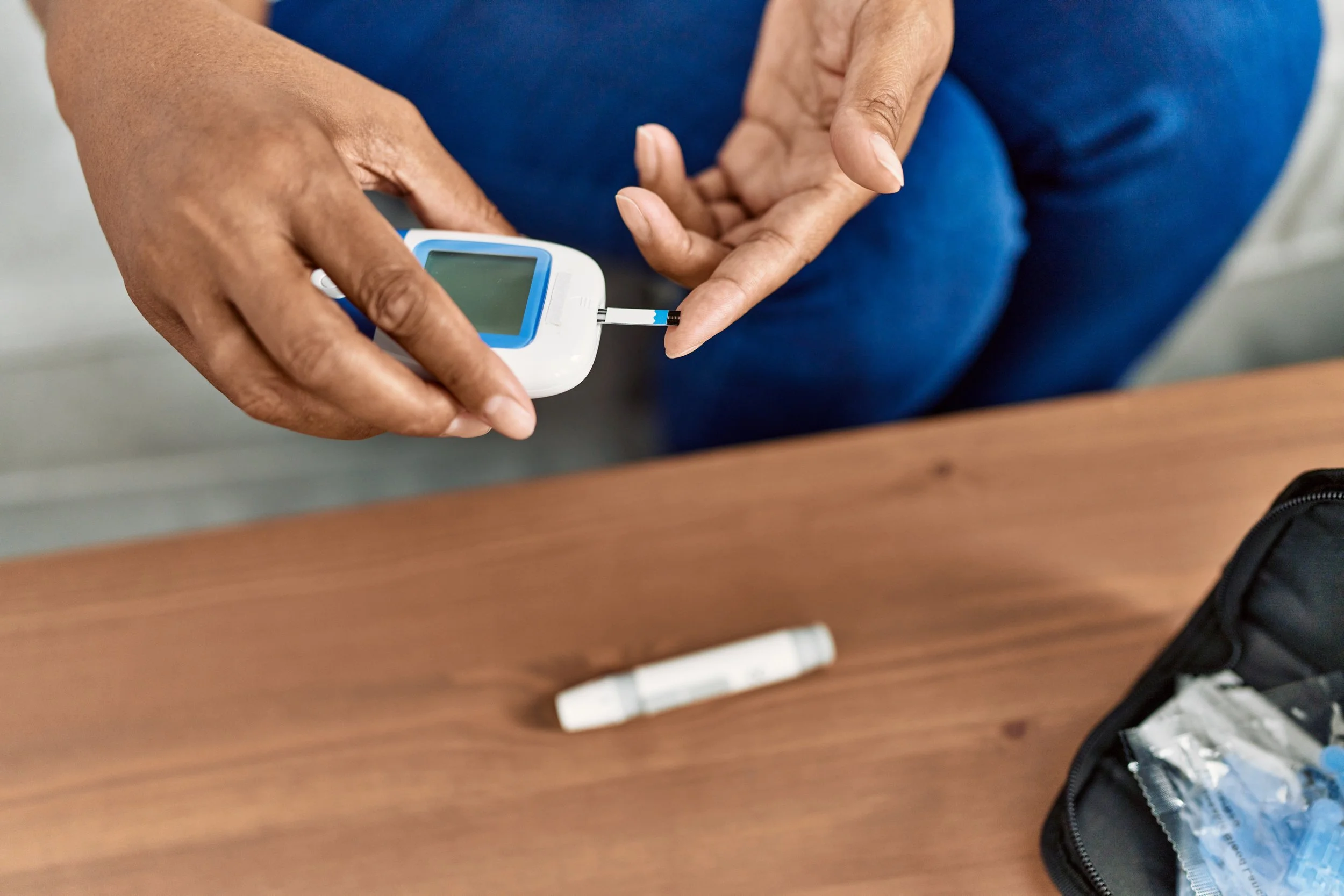 Person pricking their finger with a blood glucose meter to check blood sugar levels, with a kidney-shaped container nearby on a wooden table.