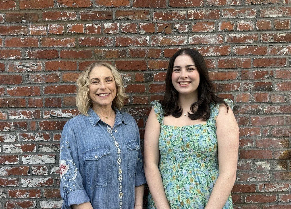 Two women smiling in front of a red brick wall, one older with blonde curly hair wearing a denim shirt, and one younger with dark straight hair wearing a floral dress.