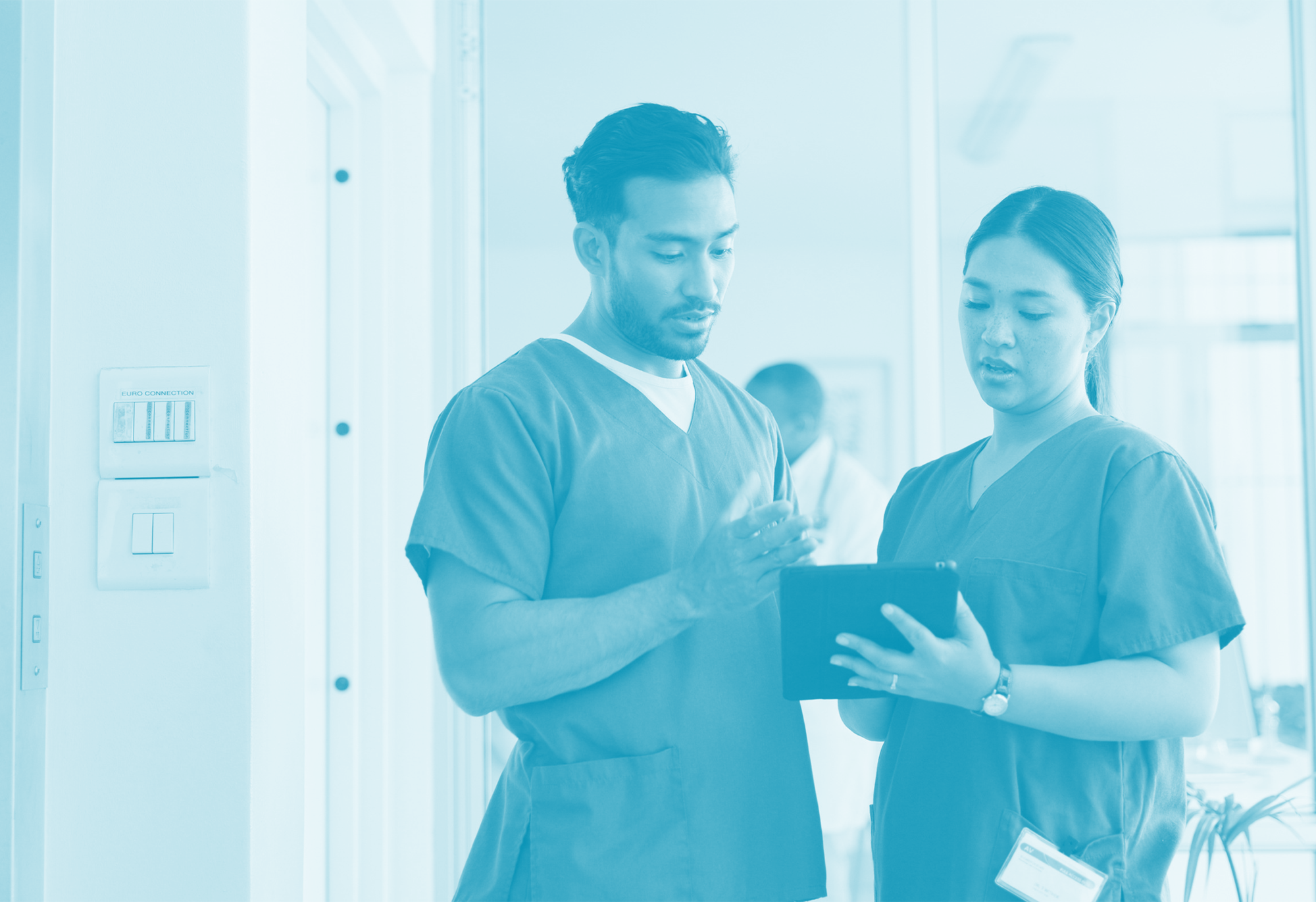 Two healthcare professionals, a man and a woman, look at a tablet together in a medical facility, with another medical worker blurred in the background.