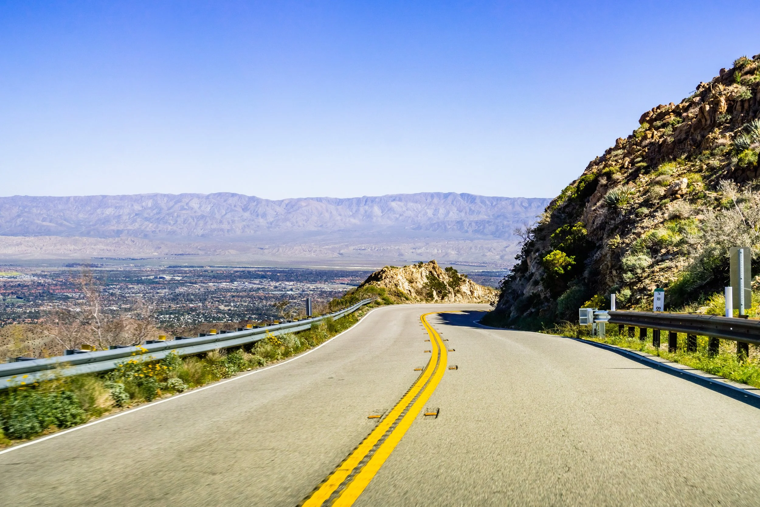 A winding mountain road with yellow dividing lines, guardrails on both sides, and a rocky hillside on the right, overlooking a valley with distant mountains under a clear blue sky.