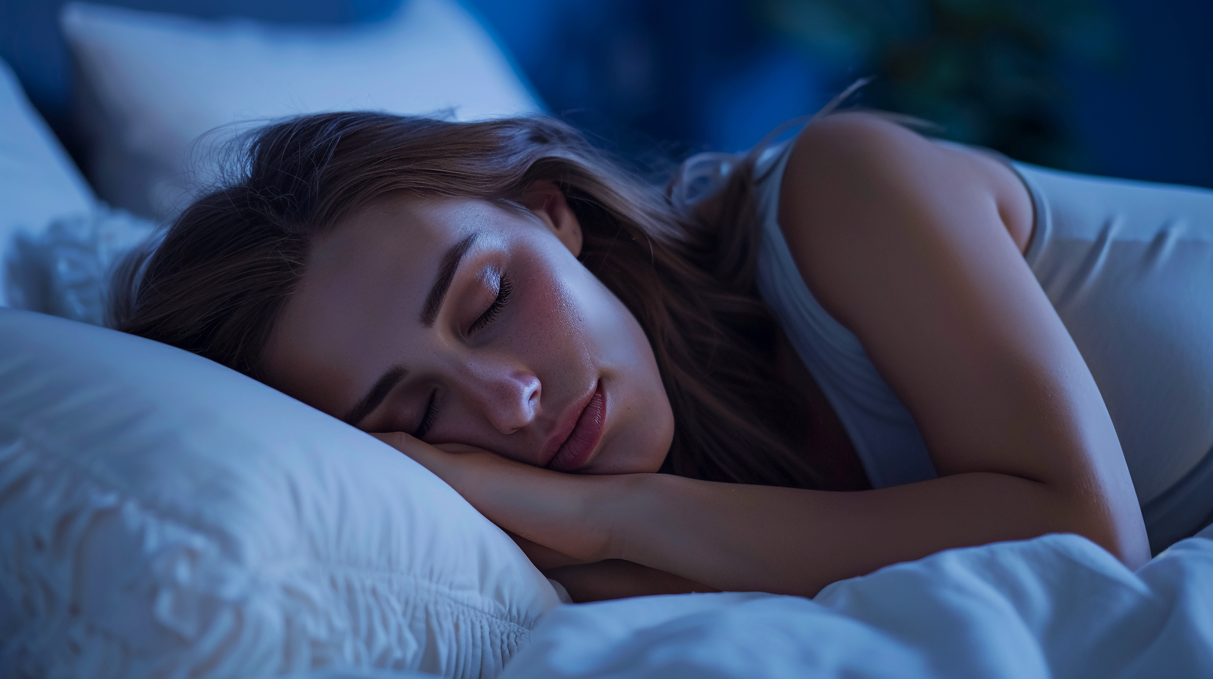 A young woman sleeping peacefully on her side in bed, with her head resting on a pillow, under soft blue light.