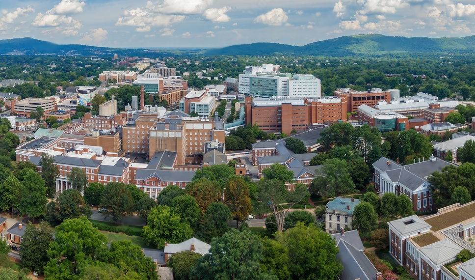 Aerial view of a university campus with multiple buildings and surrounding green trees, set against a backdrop of hills and a partly cloudy sky.