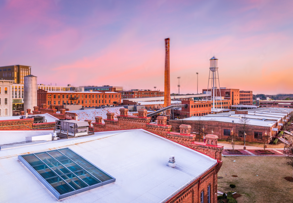 Cityscape of historic industrial buildings with brick facades, smokestack, water tower, and rooftops at sunset.