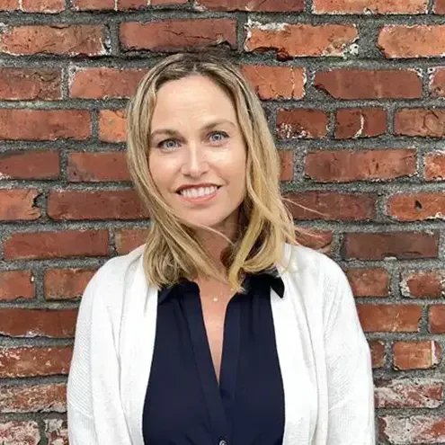 A woman with shoulder-length blonde hair, wearing a dark blouse and a light cardigan, smiling in front of a red brick wall.