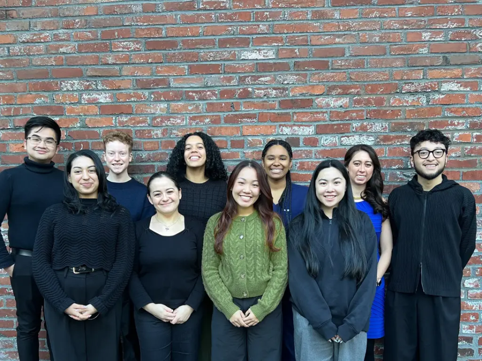 A diverse group of ten young adults standing against a brick wall, smiling for a group photo.